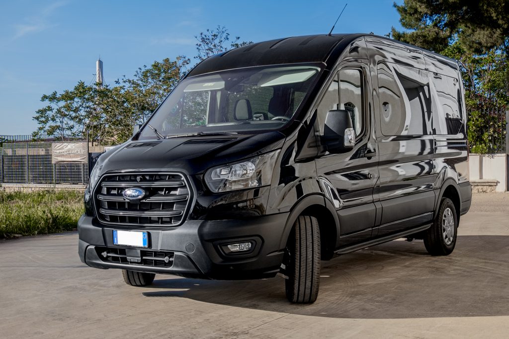 Furgone Ford Transit nero lucido con finestrini laterali, parcheggiato su piazzale in cemento vicino ad alberi e recinzione